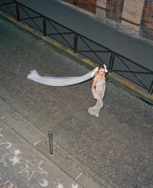 High-angle view of a woman wearing an embellished jumpsuit and veil on a cobblestone street in an urban setting at dusk.