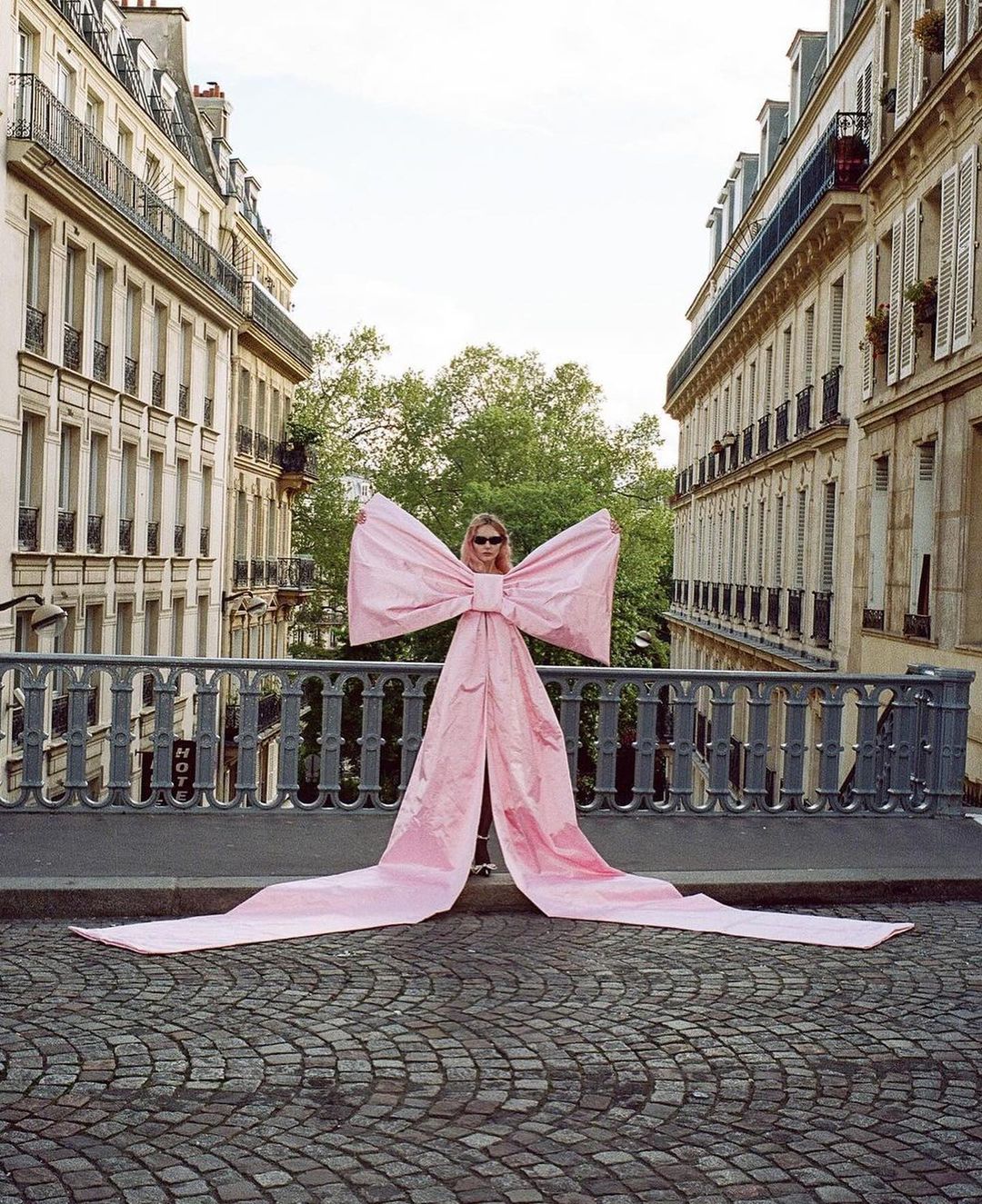 A blonde woman with sunglasses poses on a bridge in Paris, wearing a very large pink bow, with Parisian buildings in the background.
