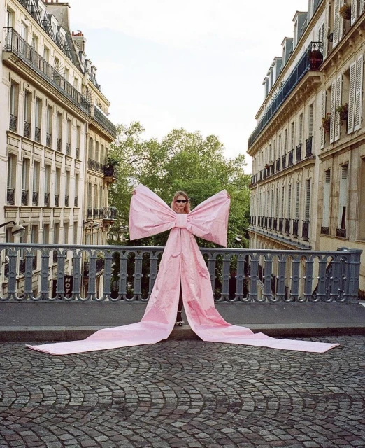A blonde woman with sunglasses poses on a bridge in Paris, wearing a very large pink bow, with Parisian buildings in the background.