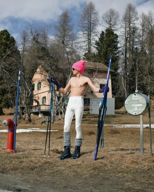 Shirtless man in pink hat poses outdoors with skis and poles near building and trees in daytime scene.