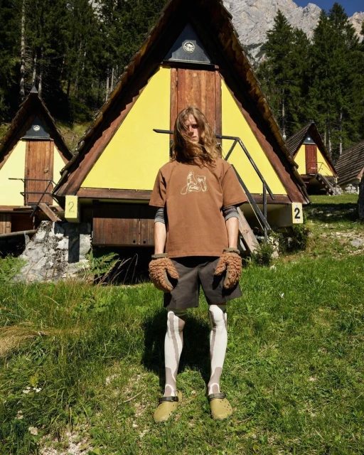 A person standing in front of yellow cabins with a mountain backdrop, wearing brown tones and unique textured gloves.