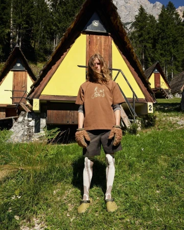 A person standing in front of yellow cabins with a mountain backdrop, wearing brown tones and unique textured gloves.