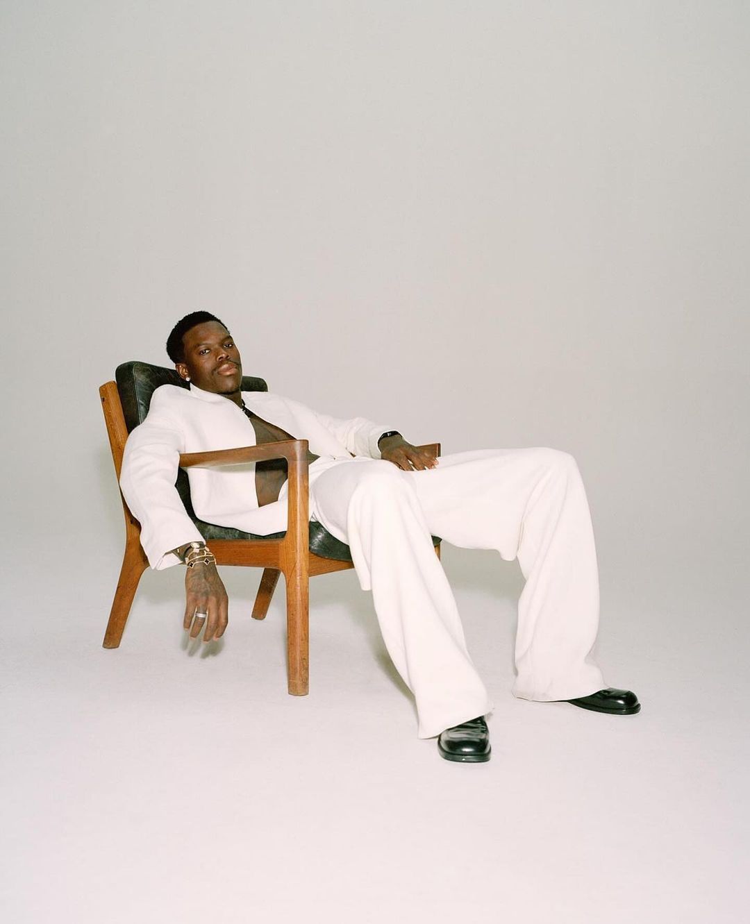 Black man in a white suit seated relaxed in a wooden armchair against a plain white studio background.