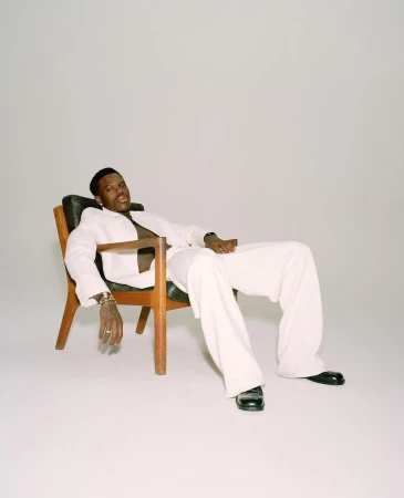 Black man in a white suit seated relaxed in a wooden armchair against a plain white studio background.