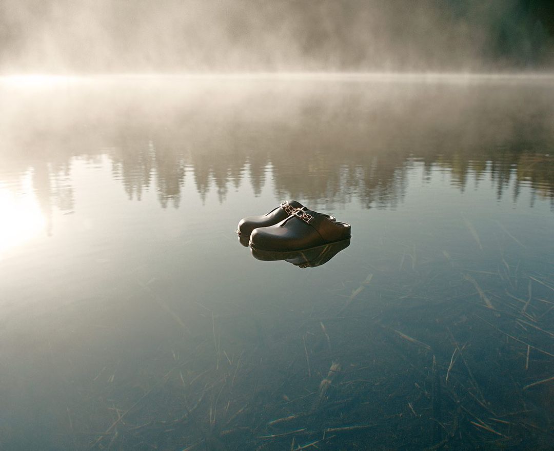 Pair of black leather clogs with gold buckle floating on a calm lake with misty forest background in the early morning light.