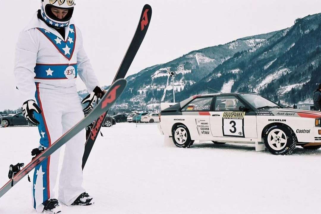Man in patriotic ski suit holds skis next to an Audi Quattro rally car on a snowy mountain, in an outdoor winter sports setting.