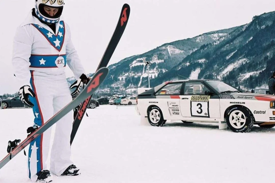 Man in patriotic ski suit holds skis next to an Audi Quattro rally car on a snowy mountain, in an outdoor winter sports setting.