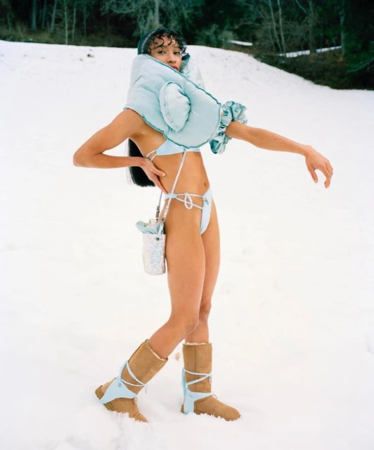 A woman in winter clothing poses in a snowy landscape, wearing a light blue bikini, tan boots with light blue accents, and a matching hood.