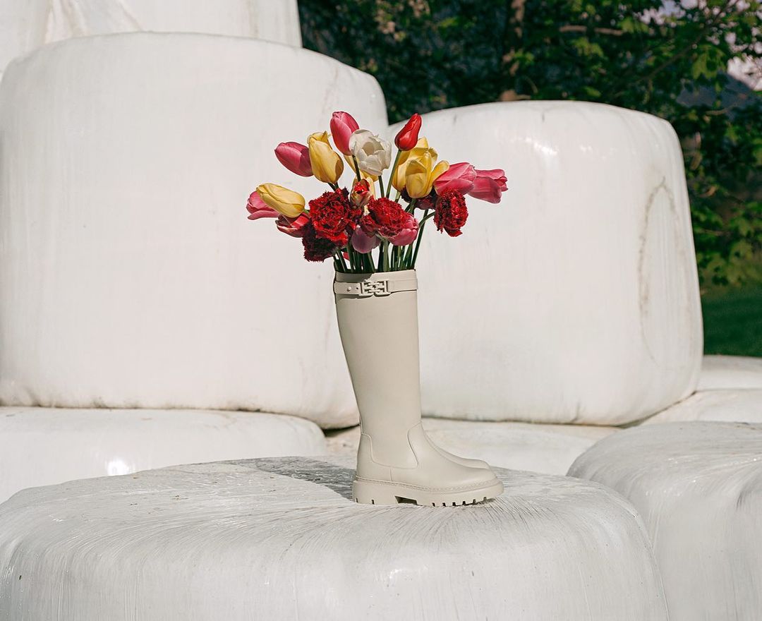 A single tan rubber boot serves as a vase filled with colorful tulips and red carnations atop a hay bale in an outdoor setting.