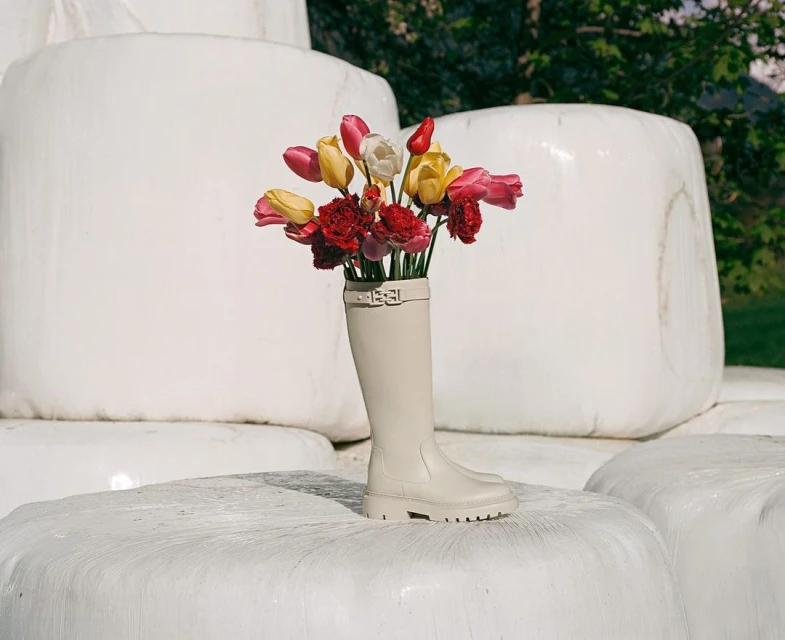 A single tan rubber boot serves as a vase filled with colorful tulips and red carnations atop a hay bale in an outdoor setting.