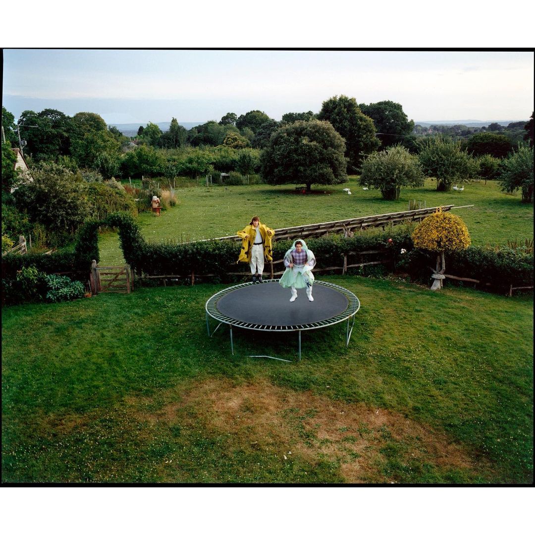Two people on a trampoline in a grassy backyard with trees and greenery under a cloudy sky, one jumping and one watching.