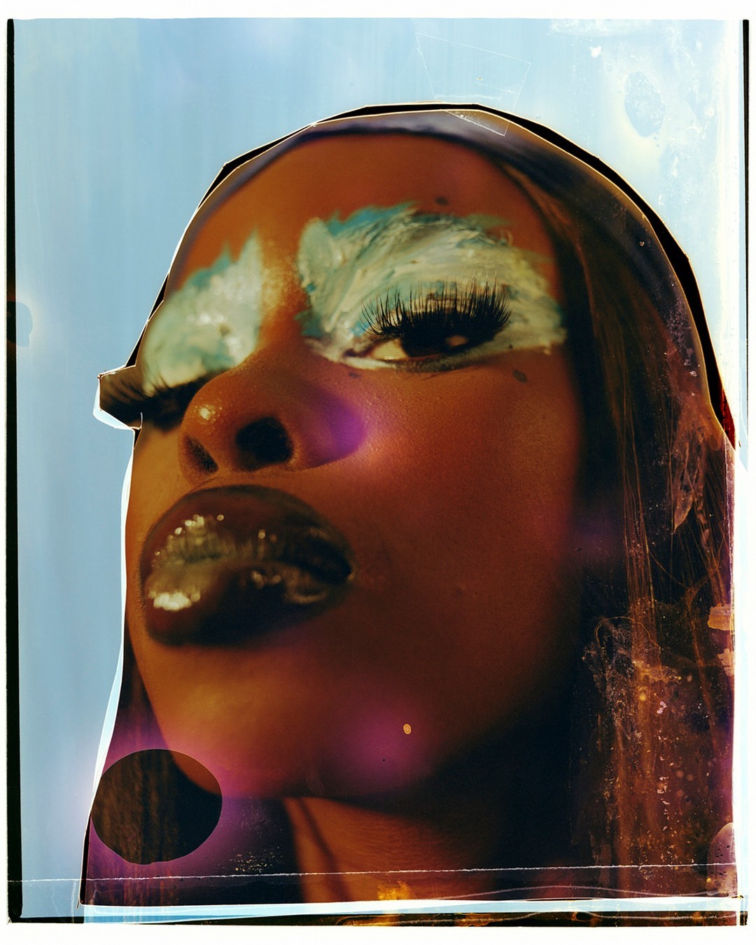 Close-up of a woman with turquoise eyeshadow and dark lipstick against a soft blue background, creating a striking and artistic makeup portrait.