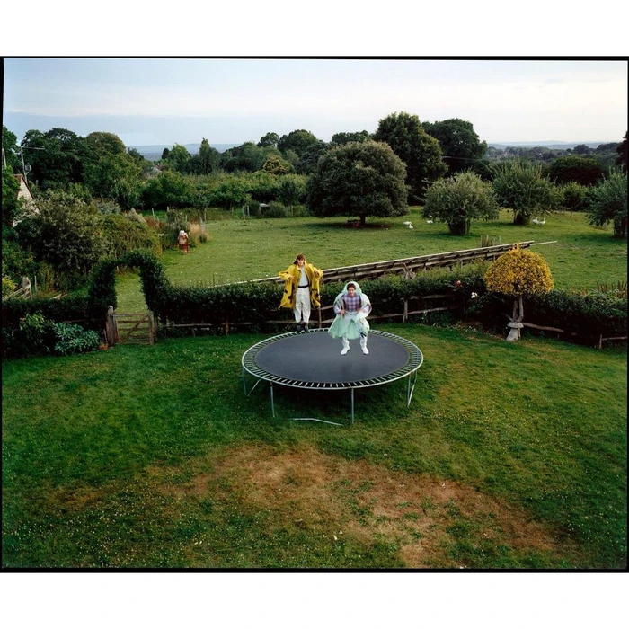 Two people jumping on a circular trampoline in a lush green lawn surrounded by trees and a hedgerow under an open sky.