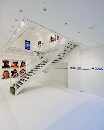 Art gallery interior featuring white walls, a metal staircase, and framed photographs on display, creating a clean and modern aesthetic.