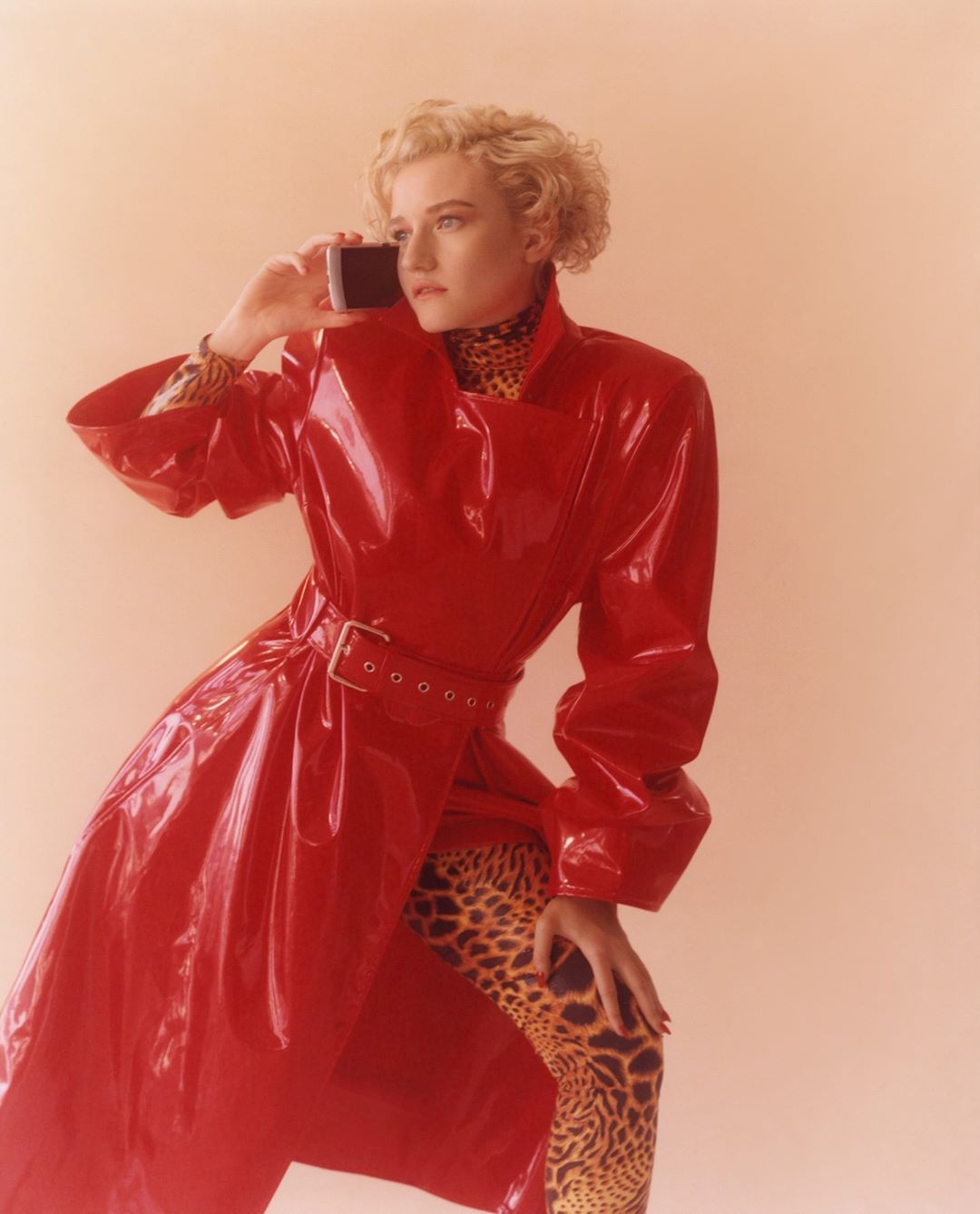 A woman poses in a red patent-leather trench coat while holding a cell phone to her ear in a studio setting with a neutral background.