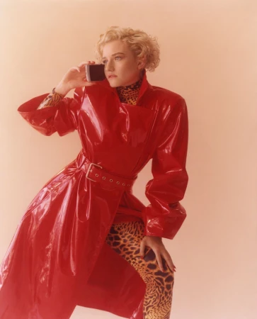 A woman poses in a red patent-leather trench coat while holding a cell phone to her ear in a studio setting with a neutral background.