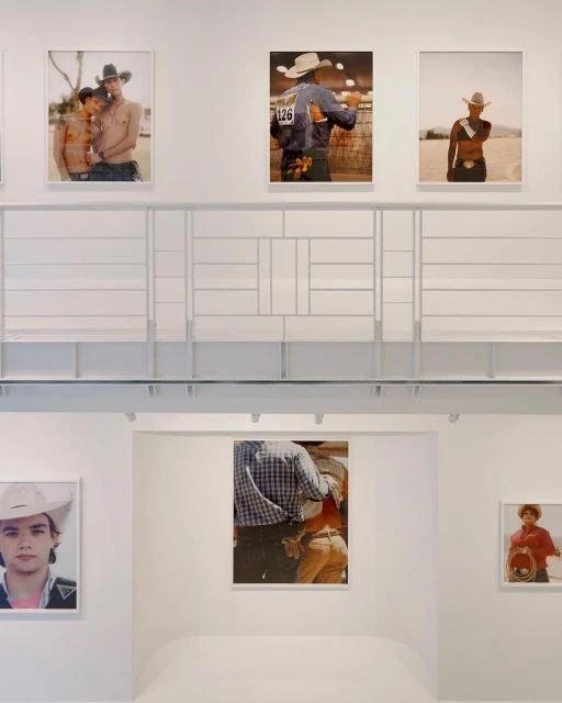 Photo gallery with framed photos of people in cowboy hats on display in a white room with a white geometric-patterned structure.