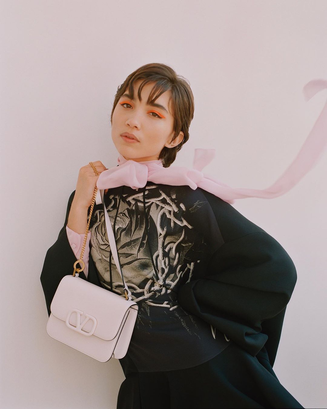 A woman with orange eyeshadow poses with a white Valentino Garavani Roman Stud top-handle bag in front of a white wall.
