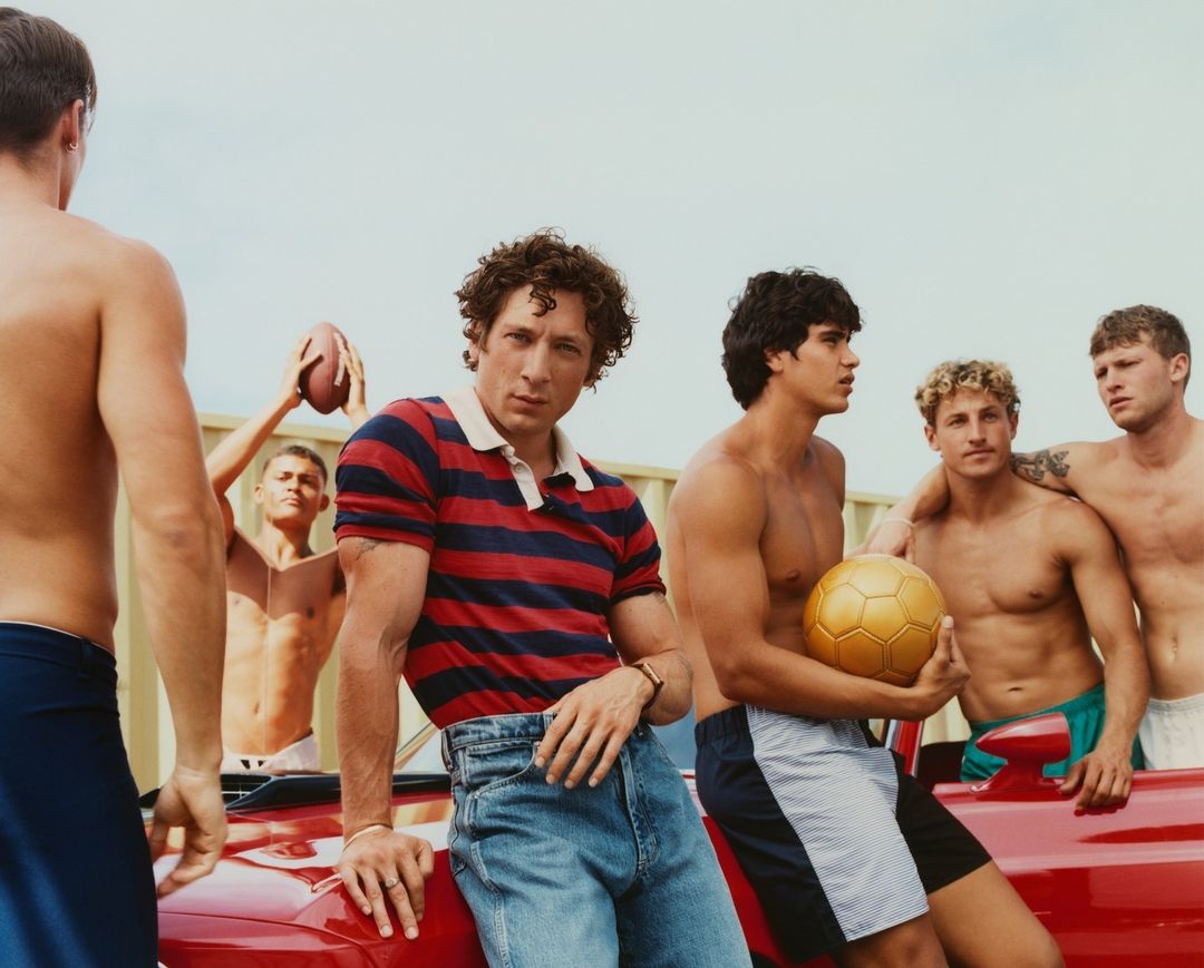 Group of young men on a red car, some shirtless, holding a soccer ball and football. Bright daylight, light sky, casual fashion.