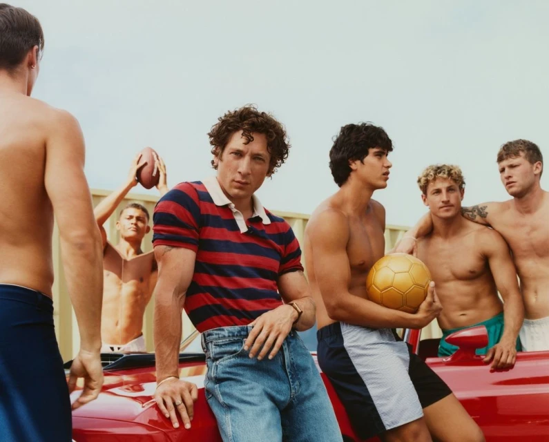 Group of young men on a red car, some shirtless, holding a soccer ball and football. Bright daylight, light sky, casual fashion.