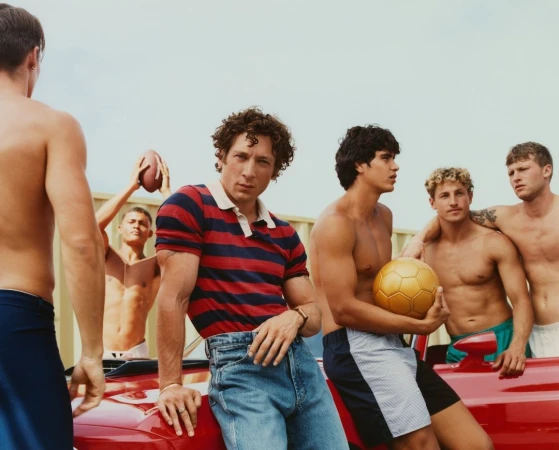 Group of young men on a red car, some shirtless, holding a soccer ball and football. Bright daylight, light sky, casual fashion.