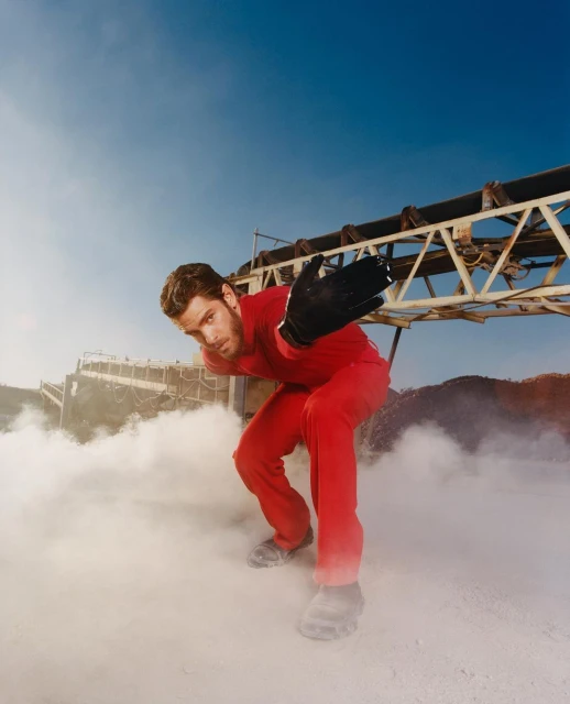 Adult male in red coveralls crouches on a dusty surface with an industrial conveyor belt in the background against a blue sky.