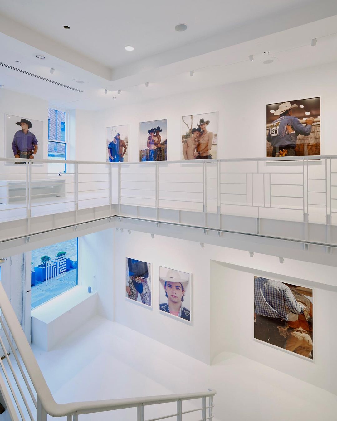 Bright, white art gallery showcases portrait photography of cowboys in hats displayed on walls with an open, multi-level layout and staircase.