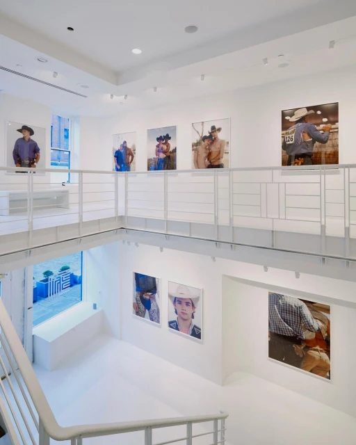 Bright, white art gallery showcases portrait photography of cowboys in hats displayed on walls with an open, multi-level layout and staircase.
