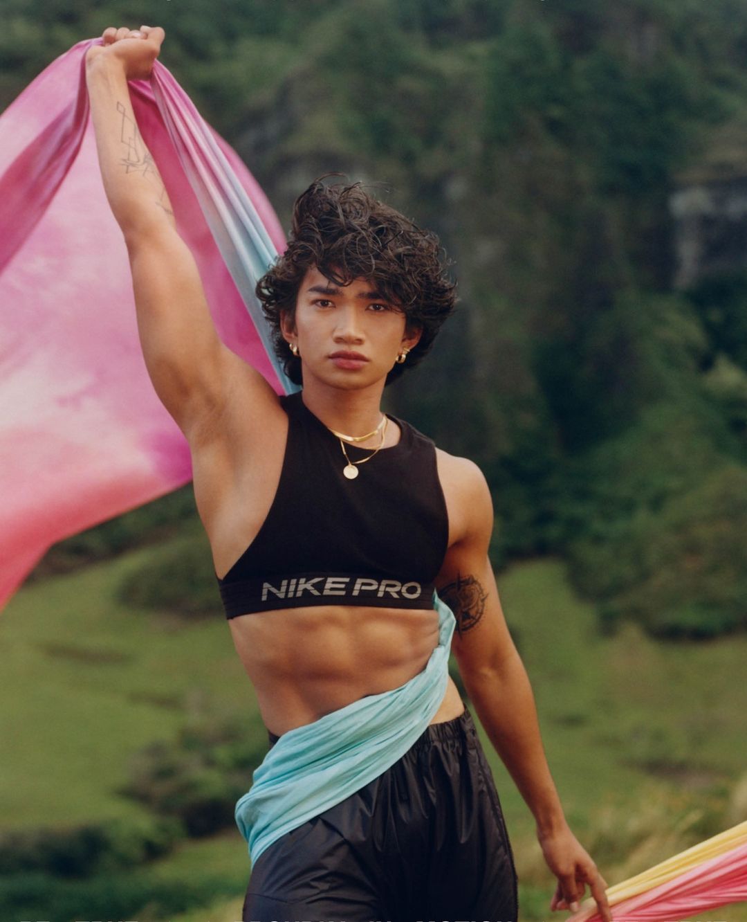Athletic adult male poses outdoors with colorful fabric, wearing a black Nike Pro sports bra and athletic shorts against a green landscape.