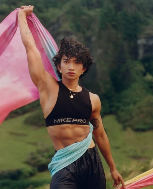 Athletic adult male poses outdoors with colorful fabric, wearing a black Nike Pro sports bra and athletic shorts against a green landscape.
