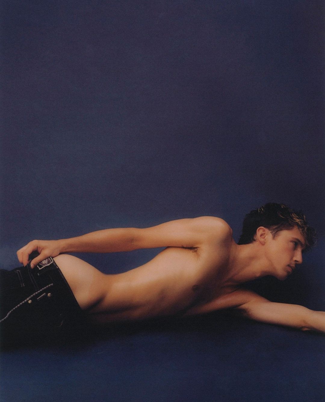 Shirtless man lying on a dark blue background wearing dark jeans photographed by Luke Gilford