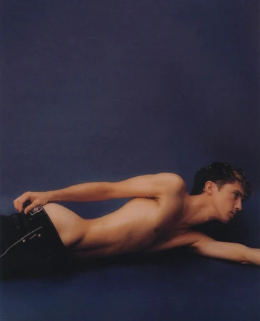 Shirtless man lying on a dark blue background wearing dark jeans photographed by Luke Gilford
