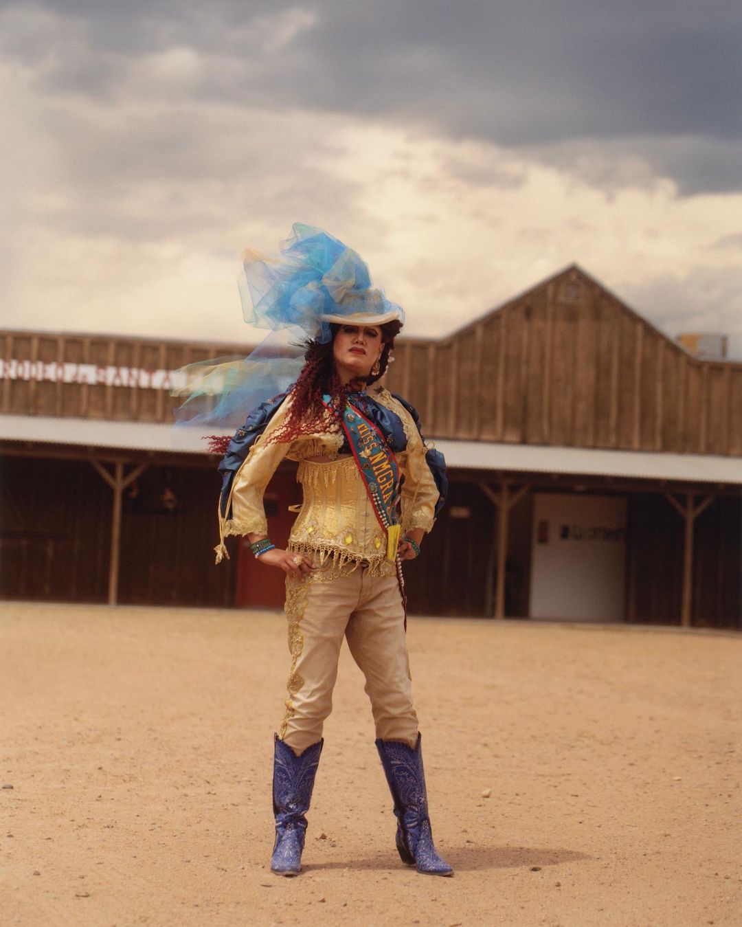 Adult female poses outdoors wearing cowboy boots, hat, and fringed attire, standing confidently in a desert-like setting under a cloudy sky.