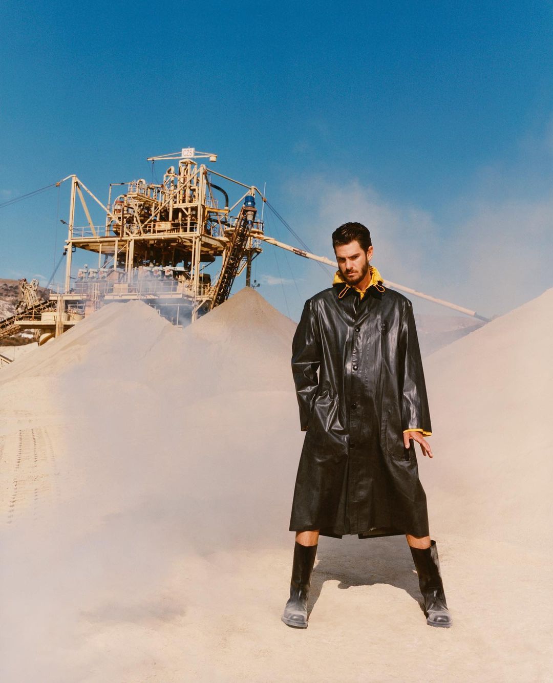 A man in a black coat and boots poses in front of industrial mining equipment under a blue sky, creating a striking fashion portrait.
