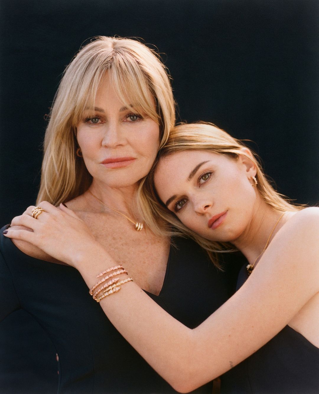 Close-up of two women, one blonde and one light brown hair, embracing each other in front of a dark background, wearing elegant dark dresses and jewelry.