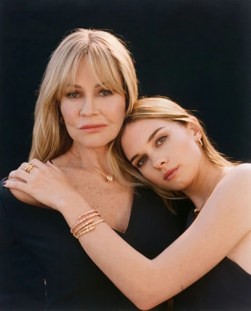 Close-up of two women, one blonde and one light brown hair, embracing each other in front of a dark background, wearing elegant dark dresses and jewelry.
