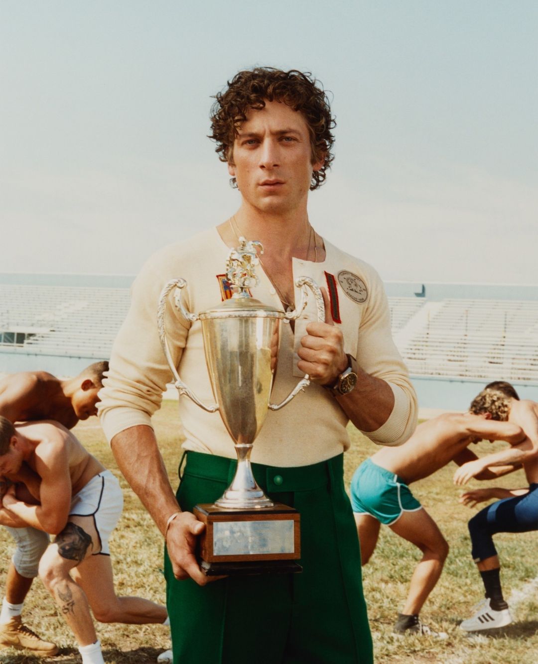 Male athlete poses with a trophy, wearing retro sportswear in an outdoor stadium, with wrestling occurring in the background.