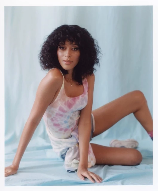 Young woman with curly black hair poses in a tie-dye tank top against a light blue backdrop, looking at the camera.