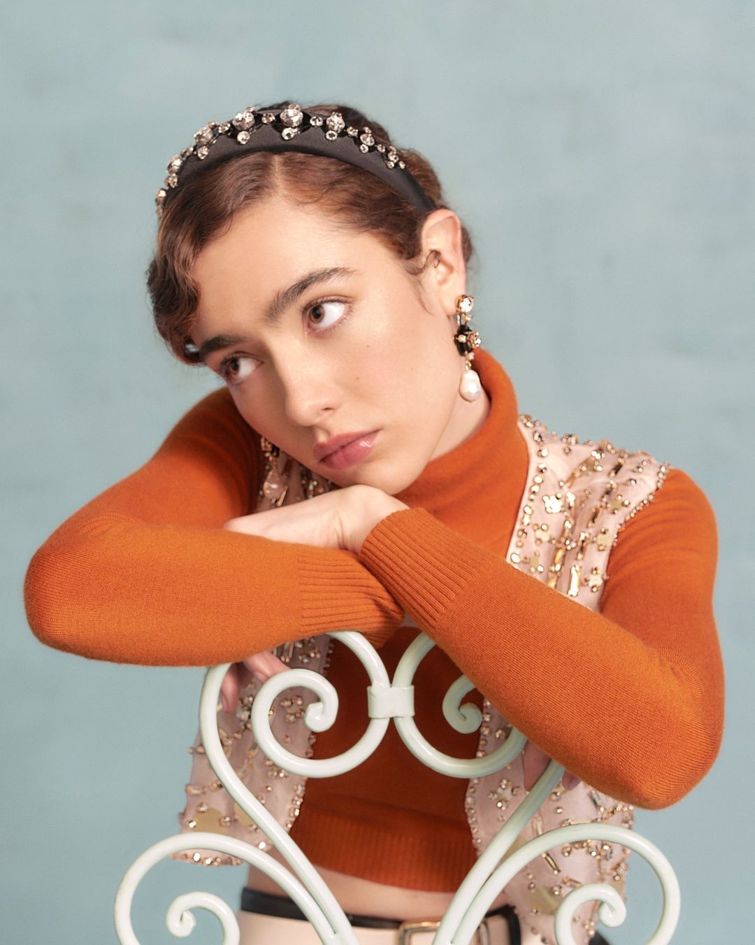 Young woman poses indoors wearing a rust-colored turtleneck, pearl earrings, and a jeweled headband, resting her arms on a chair.