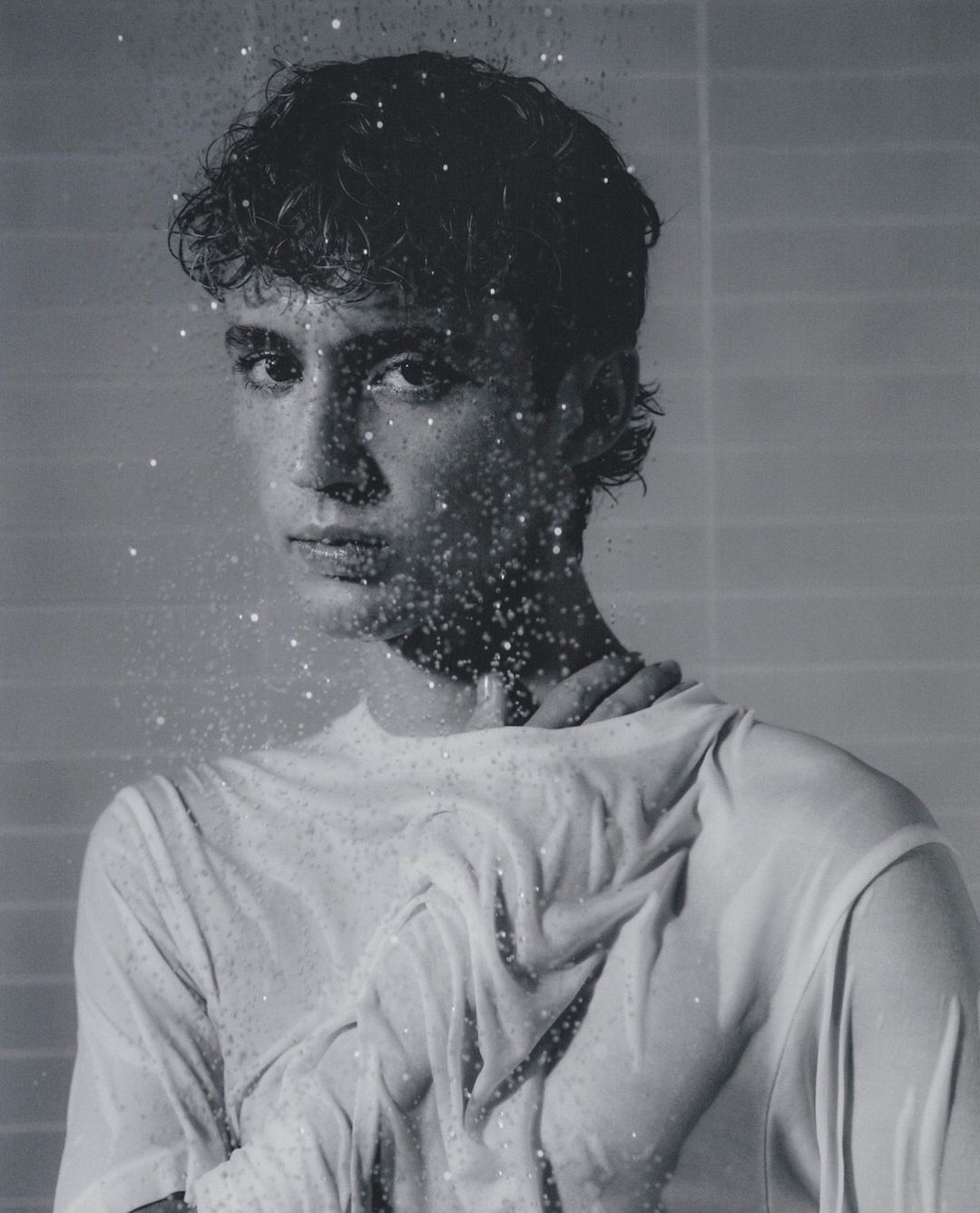 Monochromatic portrait of a young adult with wet hair and a white top, partially obscured by water droplets against a gray background.