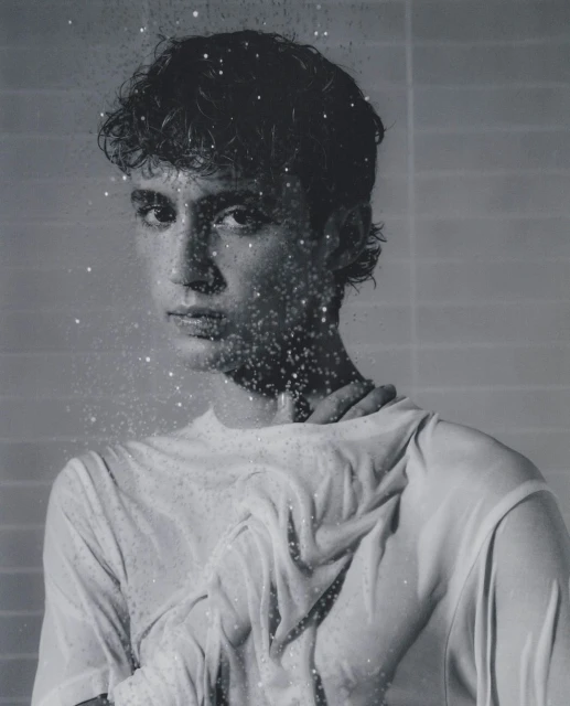 Monochromatic portrait of a young adult with wet hair and a white top, partially obscured by water droplets against a gray background.