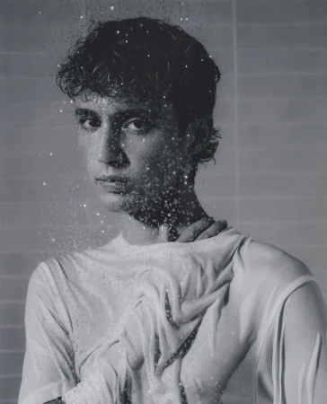 Monochromatic portrait of a young adult with wet hair and a white top, partially obscured by water droplets against a gray background.