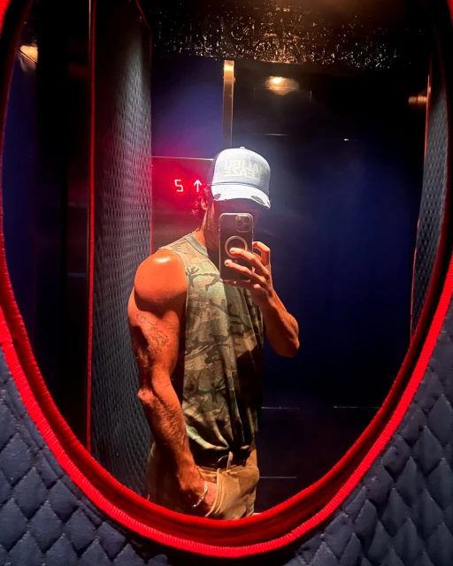 Muscular adult male takes a mirror selfie indoors. He wears a hat and camouflage tank top. The interior has blue tones and red accents.