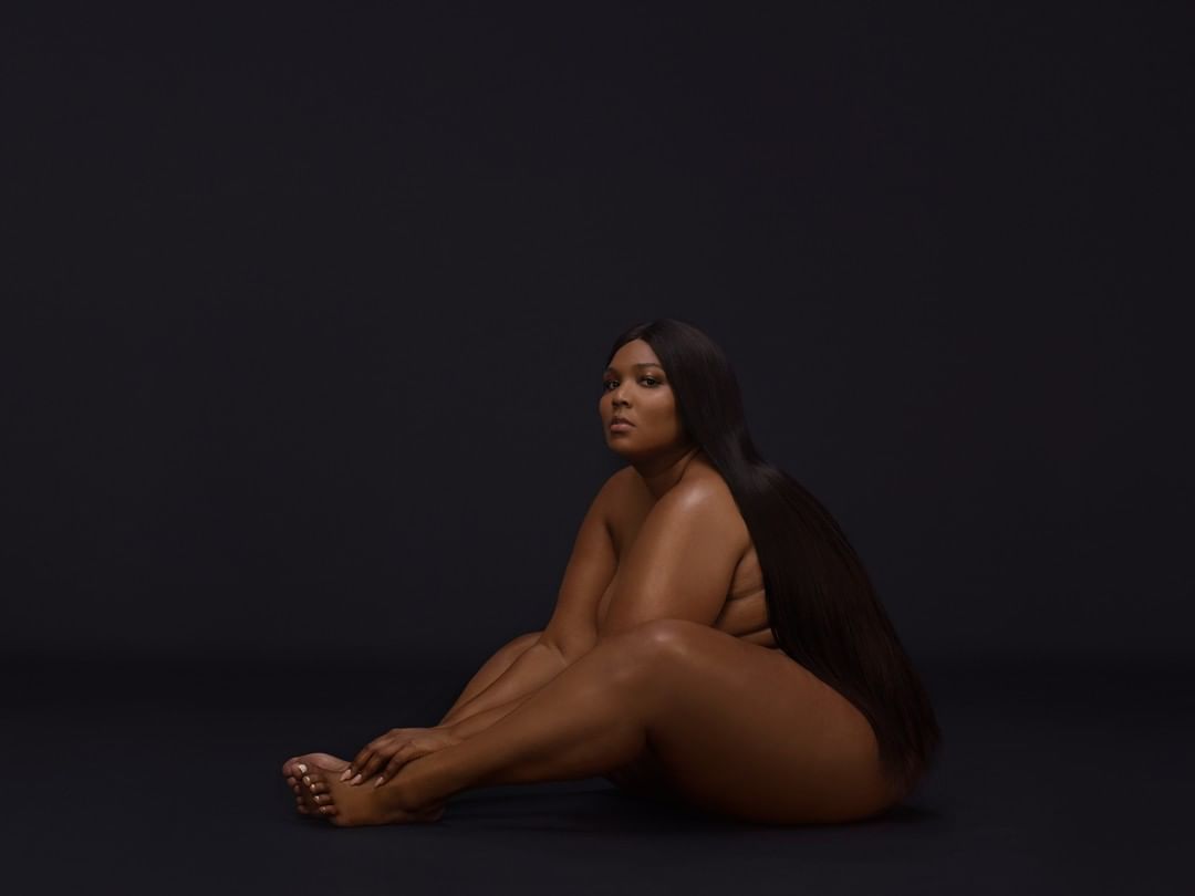 Studio portrait of Lizzo sitting on the floor against a dark background, featuring long dark hair and a thoughtful expression.