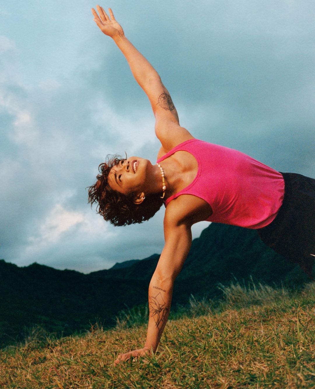 Athletic man in pink tank top stretches on grassy hillside with mountain backdrop, blue sky, and cloudy sky, outdoor workout.