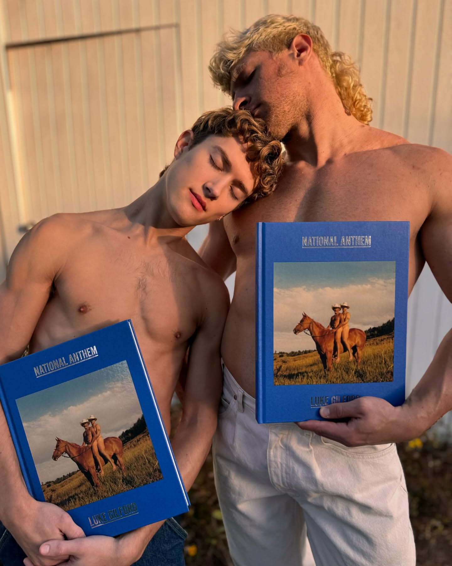 Two adult males pose shirtless outdoors holding books. One man rests his head on the other's head, with natural light and neutral background.