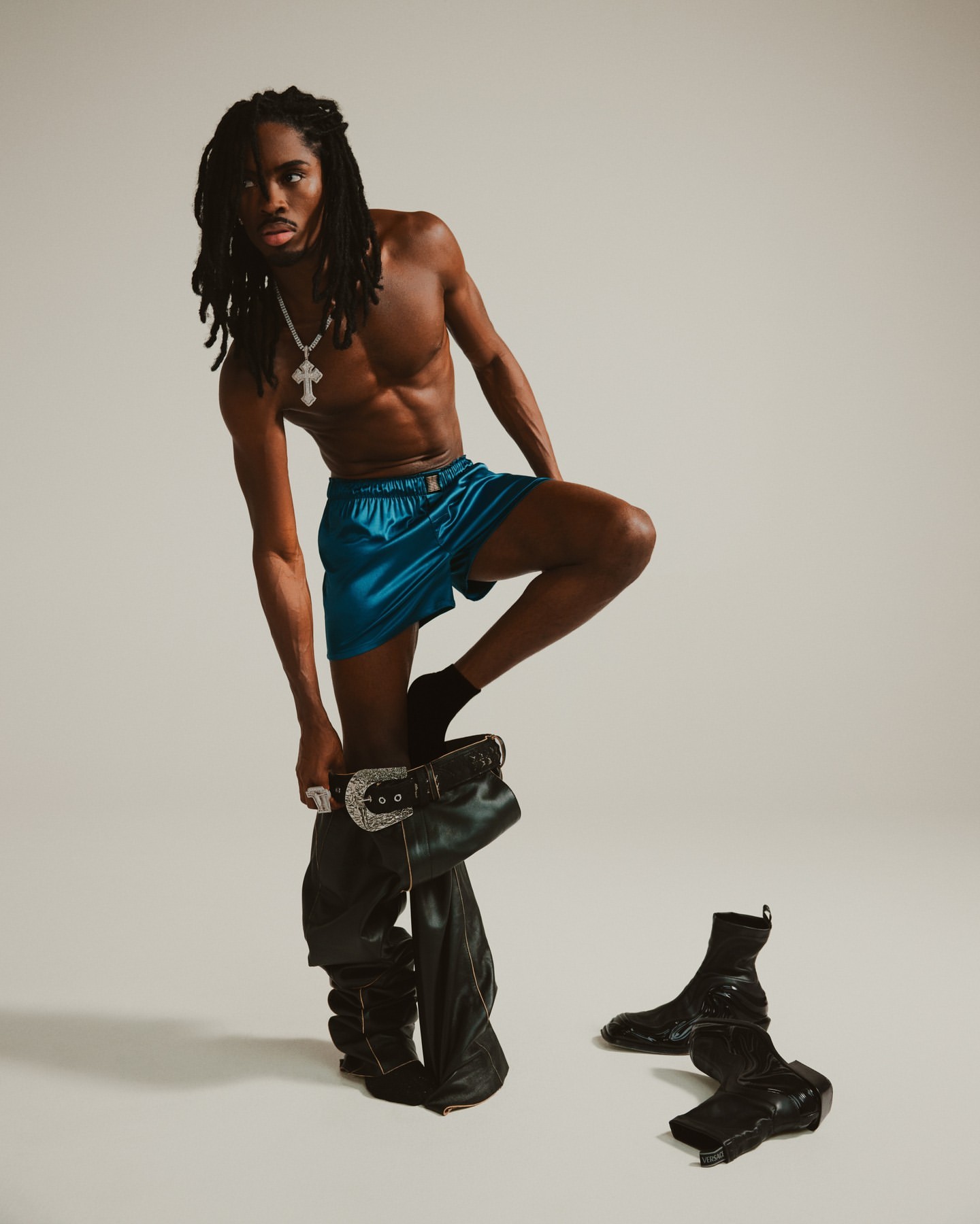 Shirtless Black male model with dreadlocks wears blue shorts and leather cowboy boots in a studio fashion shot.