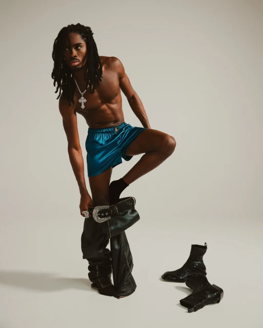 Shirtless Black male model with dreadlocks wears blue shorts and leather cowboy boots in a studio fashion shot.
