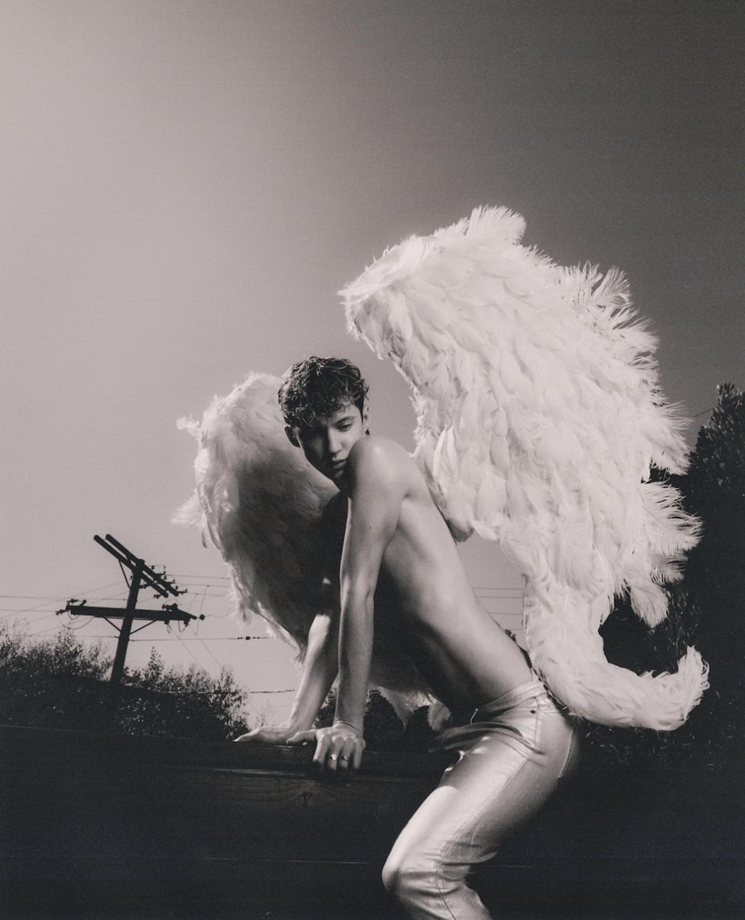 Monochrome portrait of adult male with feathered angel wings in silver pants leaning on a ledge against an outdoor background sky and trees.