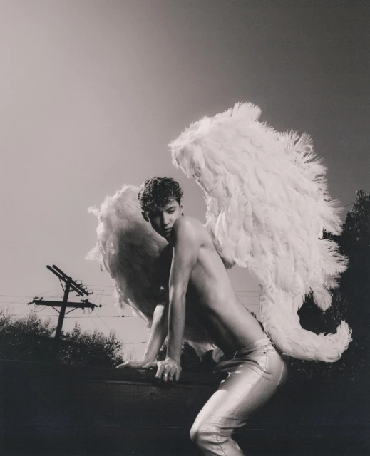 Monochrome portrait of adult male with feathered angel wings in silver pants leaning on a ledge against an outdoor background sky and trees.